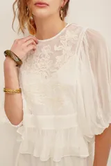 Camisa blanca de manga larga abullonada, con cuerpo de red transparente con bordados florales en tono crudo y aplique de flor grande fucsia en el hombro.