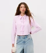 Camisa cropped de manga larga en tejido tricoline, con estampado de rayas verticales finas en color rosa y blanco. Presenta cuello camisero tradicional y cierre frontal decorado con tres lazos de tela. Diseño de corte corto y ajuste recto.