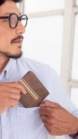 Billetera de cuero sintético color marrón oscuro con textura granulada, presenta una banda elástica decorativa central con rayas blancas y tostadas. Cuenta con compartimentos internos para billetes, tarjetas y visor.