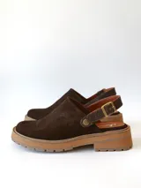 Zueco de cuero gamuzado color chocolate, con diseño de punta cerrada y tira ajustable con hebilla metálica en el talón. Presenta una suela de plataforma dentada en tono marrón claro.