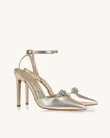 Zapato de tacón alto color dorado con pulsera al tobillo y detalle de moño plateado con brillos en la punta.
