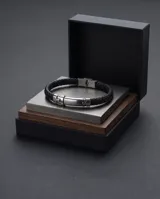 Pulsera de cuero trenzado negro con placa central de acero inoxidable y cierre de seguridad metálico.