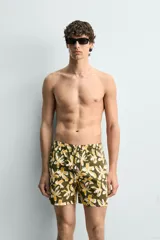 Bañador de hombre con estampado floral en tonos amarillo y blanco sobre fondo verde oliva. Presenta cintura elástica ajustable con cordón, bolsillos frontales y forro interior.