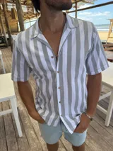 Camisa de manga corta con cuello cubano, confeccionada en algodón con diseño de rayas verticales en tonos gris y blanco. Presenta cierre frontal con botones de madera.
