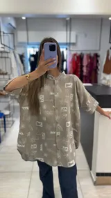 Camisa oversize de manga corta, color beige, con cuello mao y botones frontales. Presenta un estampado repetitivo en blanco con motivos playeros como palmeras, tablas de surf y olas.