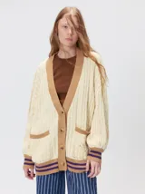 Cardigan de punto trenzado en color negro con detalles en color crema en los bolsillos, puños y ruedo. Presenta un diseño oversize, cierre frontal con botones y bordado decorativo en uno de los bolsillos delanteros.