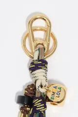 Llavero de diseño náutico compuesto por cabos trenzados en tonos beige, marrón y violeta, con herrajes metálicos dorados y dije cuadrado con logo de la marca esmaltado.