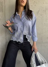 Camisa celeste a rayas verticales blancas, con cuello clásico, cierre frontal con botones y mangas largas. Presenta detalle de bordado calado con motivos florales en el ruedo.