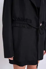 Blazer negro con solapas de muesca, corte holgado, cierre frontal con lazo y detalle de volados en los bolsillos.