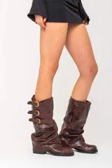 Botas de caña alta confeccionadas en cuero vacuno con acabado brillante. Presentan un diseño de caña holgada con tres correas horizontales ajustables mediante hebillas metálicas doradas. Cuentan con un taco bajo cuadrado y suela de goma resistente.