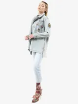 Chaqueta de denim en tono azul claro con efecto lavado, corte holgado y cierre frontal con botones. Presenta múltiples insignias decorativas, incluyendo broches con cristales, una insignia con cinta y detalles de jacquard en los laterales. Cuenta con bolsillos frontales y aberturas laterales.