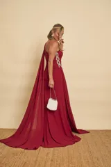 Vestido largo de fiesta color bordó, de un solo hombro, con detalles de pedrería en el tirante y fruncidos laterales. Cuenta con tajo pronunciado en la pierna y una capa larga vaporosa que cae desde el hombro.