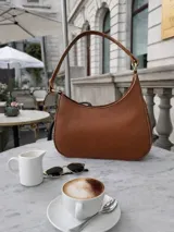 Cartera tipo hobo de cuero con textura granulada, diseño de media luna y asa fija para llevar al hombro.