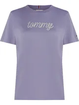 Remera de manga corta, cuello redondo, color lila/malva, con el logo "Tommy" escrito en cursiva con apliques de strass o pedrería. Presenta un pequeño logo de bandera de Tommy Hilfiger en la manga izquierda.