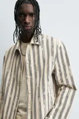 Cazadora de hombre de corte relajado, color beige claro con rayas verticales en azul oscuro con efecto desgastado o estampado. Presenta cuello solapa, manga larga, bolsillos de vivo en cadera y cierre frontal con botones a presión.