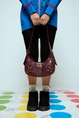 Cartera de hombro estilo hobo en color bordó, confeccionada en cuero vegano con acabado texturizado. Presenta una hebilla decorativa central, herrajes metálicos plateados, correas laterales con hebillas y una correa de hombro regulable.