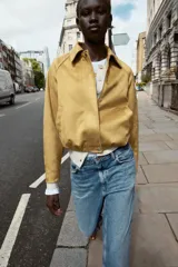 Chaqueta bomber de efecto ante en color tostado, con cuello solapa, manga larga y bolsillos laterales. Presenta un diseño con bajo acabado en pliegues que aporta volumen y cierre frontal con cremallera y botones a presión.