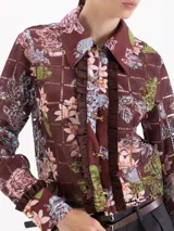 Blusa de manga larga con estampado floral y geométrico en tonos marrones, con cuello clásico, cierre frontal con ganchos metálicos y detalle de volados laterales en la tapeta.