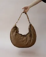 Bolso tipo hobo de gran tamaño, confeccionado en gamuza color marrón oscuro. Presenta un diseño artesanal con costuras visibles y detalles de tientos en el cuerpo, asa integrada con pespuntes a mano y cierre superior.