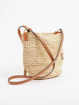 Bandolera de rafia natural de forma redondeada, con correa larga fina de cuero marrón, cierre superior y detalle de aplique en cuero con la letra K.