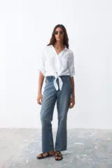 Camisa blanca de lino con cuello clásico, mangas 3/4 con botón para enrollar, bolsillo en el pecho y cierre frontal con botones. Se ata en la cintura.