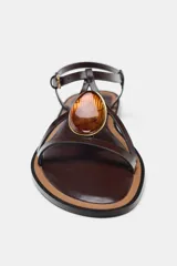 Sandalia plana de cuero color marrón oscuro (bordó/caoba) con tira en T y hebilla ajustable en el tobillo. Presenta un aplique decorativo curvo en tono ámbar/marrón rojizo con borde dorado en la tira del empeine.