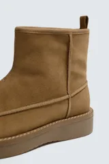 Bota de caña corta fabricada en piel con acabado serraje, con detalle de costura en el empeine, forro interior de pelo sintético y suela plana a tono.