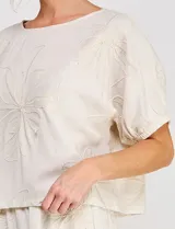 Blusa corta de lino y viscosa color beige, con bordado de grandes flores al tono, cuello redondo, mangas cortas abullonadas y corte holgado.