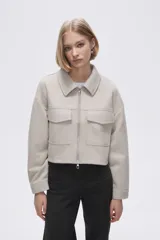 Chaqueta de eco-cuero en color beige, con diseño holgado, cuello tipo camisa y cierre frontal metálico. Presenta dos bolsillos frontales con solapa y mangas largas con puños rectos.