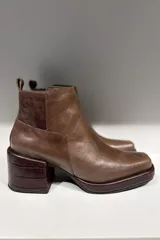 Bota corta de cuero color marrón, con diseño de punta cuadrada y taco cuadrado con textura grabada. Presenta costuras visibles y una lengüeta en la parte posterior.