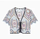 Remera corta de poliéster color crudo con bordados en tonos azules, marrones y morados, escote en V con ribete oscuro y cierre frontal con cordones y cuentas.