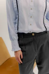 Camisa de algodón blanca con cuello a la base, cierre frontal con botones y mangas largas con puños delicados. Presenta volados verticales y detalles de cintas de terciopelo en contraste a lo largo del frente.