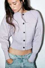 Chaqueta corta de punto color lila, con cuello redondo, manga larga con puños acanalados y cierre frontal con botones negros. Presenta un diseño con acabado en rib en el ruedo.