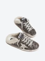 Zuecos Golden Goose modelo Super-Star Sabot, con diseño de zapatilla sin talón. Presentan exterior cubierto de glitter negro y dorado, estrella característica en el lateral, cordones y forro interior de shearling sintético.