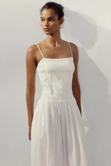 Vestido midi de color blanco con escote recto y tirantes finos ajustables. Presenta un cuerpo con tejido elástico tipo nido de abeja y detalles de flores bordadas en la parte superior. Cuenta con bolsillos laterales ocultos y forro interior.