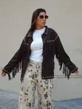 Chaqueta de gamuza color chocolate, con diseño de flecos en el frente y la espalda, decorada con tachas metálicas en cuello, hombros y puños. Cuenta con cierre frontal de botones y forro interior.