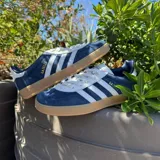 Championes adidas Gazelle de diseño clásico, confeccionados en gamuza azul con las icónicas tres tiras laterales en blanco. Presentan detalles en blanco en el talón y la lengüeta, con el nombre del modelo en dorado y una suela de goma color caramelo.