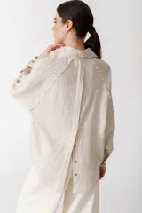 Camisa color ladrillo con rayas verticales finas color crema, de corte recto y mangas largas con puños abotonados. Presenta cuello camisero, cierre frontal con botones y detalle de flecos en los hombros.