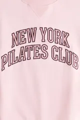 Buzo oversize rosado con interior cepillado suave, cuello redondo, hombros caídos y estampa frontal con la inscripción 'New York Pilates Club'. Cuenta con ribetes acanalados en el cuello, los puños y el bajo.