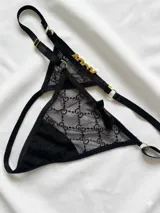 Tanga de tul translúcido negro con diseño de encaje tipo monograma, tiras elásticas regulables con argollas doradas y detalle de letras en strass formando la palabra "TODA".