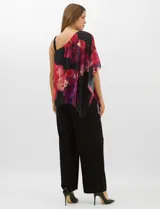 Enterito de seda fría con pantalón corte palazzo de pierna ancha. Presenta un diseño de un hombro descubierto con tirante fino y una sobrecapa asimétrica de gasa estampada con motivos florales en tonos rojos y fucsia sobre fondo negro que cae desde el hombro derecho.