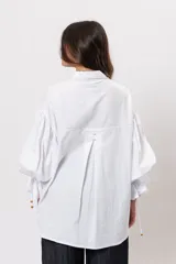 Camisa de algodón blanca de silueta amplia, con cuello camisero, botonadura frontal con botones decorativos, mangas abullonadas extragrandes y detalles de tiritas ajustables con cuentas de resina en los puños.