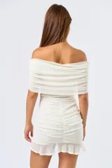Vestido corto de mesh color blanco, con diseño off-shoulder y escote con volados. Presenta un ajuste drapeado y fruncido en la parte central de la falda, finalizando con un ruedo con volado sutil.