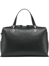 Cartera tipo satchel de Calvin Klein en color negro, confeccionada en material sintético con textura granulada. Presenta doble asa de mano, correa de hombro ajustable y extraíble, y cierre superior con cremallera. Incluye accesorios colgantes decorativos: un monedero circular y un pequeño estuche con solapa, ambos con el monograma de la marca.