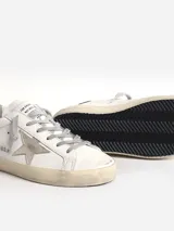 Zapatillas urbanas Golden Goose modelo Super-Star, de cuero blanco con estrella plateada en el lateral, detalle de talón en cuero negro y suela de goma color crema con efecto desgastado.