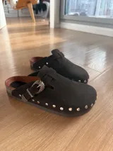 Zueco de gamuza marrón oscuro, forrado en cuero vacuno, con tira ajustable con hebilla metálica decorativa y tachas plateadas alrededor de la suela.