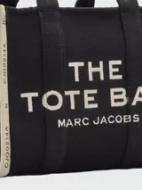 Bolso tipo tote bag de Marc Jacobs en color beige, confeccionado en lona resistente. Presenta el texto "THE TOTE BAG" y "MARC JACOBS" impreso en la parte frontal en color negro. Cuenta con asas superiores y una correa de hombro ajustable y extraíble, además de ribetes laterales negros con inscripciones.