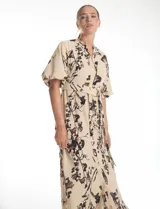 Vestido maxi de crepe color beige con estampado floral en tonos marrones y negros. Presenta cuello a la base, escote en V, abertura central con botones y cintura con lazo de ajuste y hebilla.