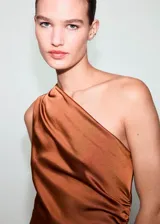 Vestido midi de satén color cobre con diseño asimétrico de un solo hombro. Presenta un detalle fruncido lateral que recorre el costado, cierre de cremallera lateral y una abertura en el bajo.