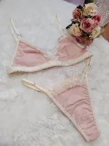 Conjunto de lencería compuesto por un bralette triangular y una tanga, confeccionados en algodón color rosado con detalles de encaje en tono crema. El bralette cuenta con breteles ajustables y el conjunto presenta terminaciones delicadas.